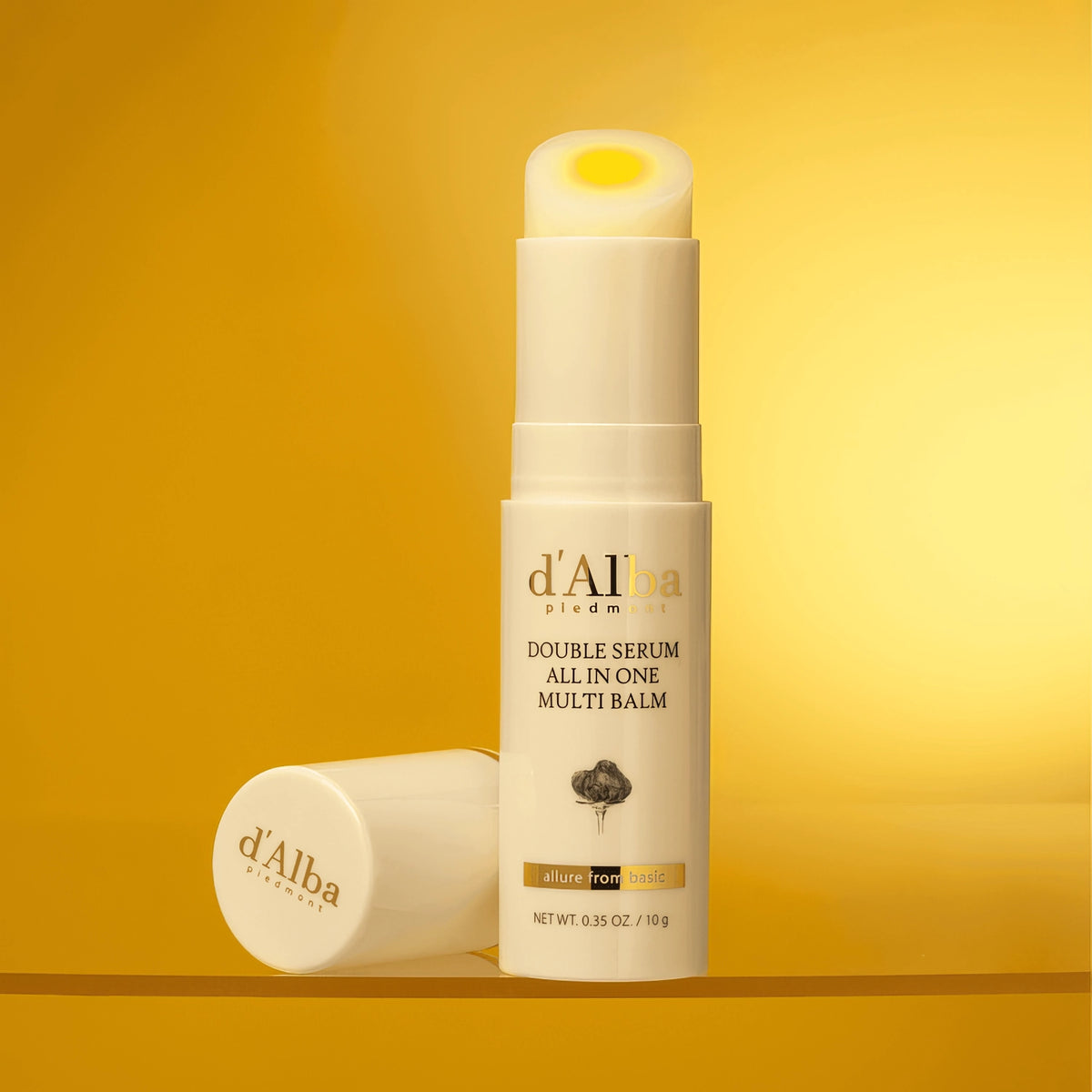 d'Alba Double Serum All In One Multi Balm 10g – kleiner goldfarbener Stick vor goldenem Hintergrund mit Blüten